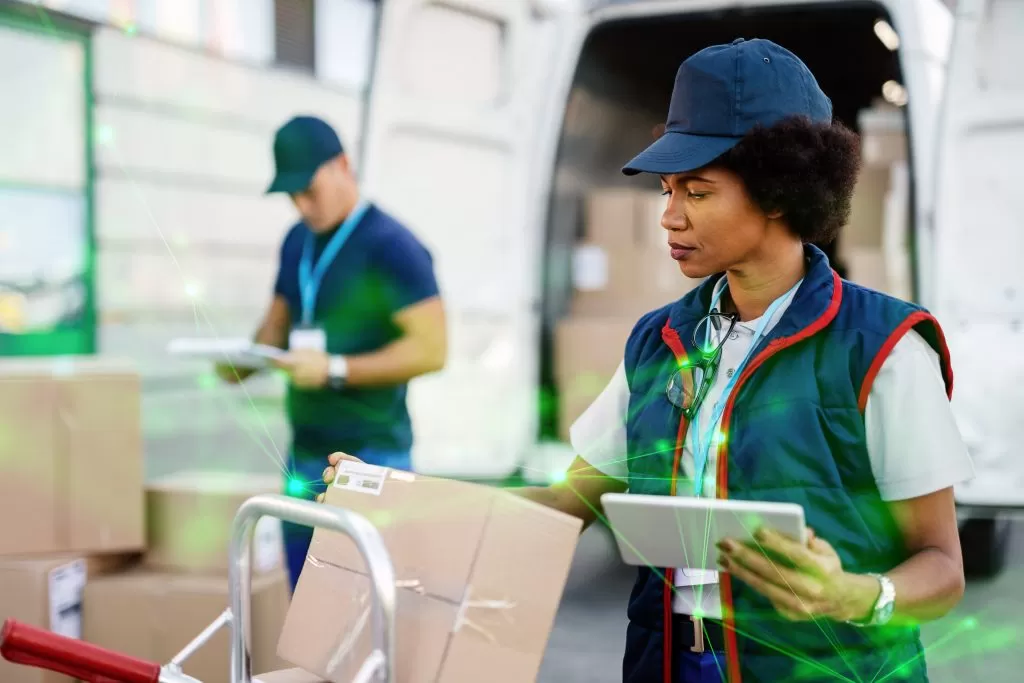 female worker using digital tabelt while doing package inventory befire delivery her coworker is background with green plexus